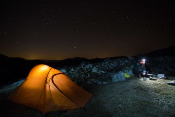 Hiker Dan Dinu heating up water beside his tent under starry sky over a rocky limestone ridge in Mehedinti plateau geopark, Romania